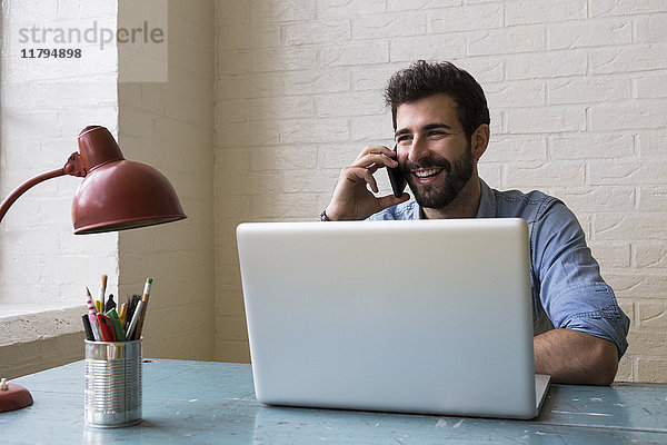 Lächelnder Mann am Telefon sitzt am Schreibtisch in seinem Loft.