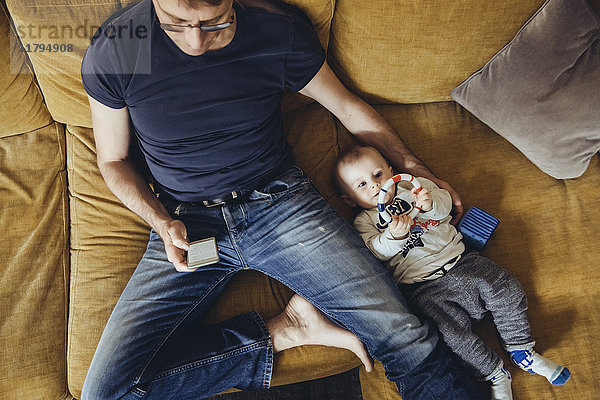 Kleiner Junge  der neben seinem Vater auf der Couch liegt und ein Smartphone benutzt.