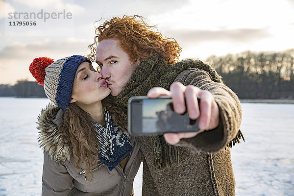 Ein Paar  das sich auf dem zugefrorenen See küsst und einen Selfie nimmt.