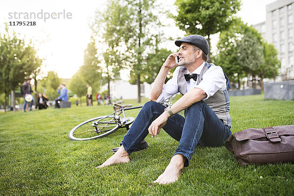 Selbstbewusster Geschäftsmann mit Fahrrad und Smartphone im Stadtpark auf Rasen sitzend