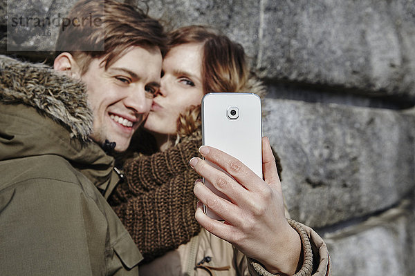 Glückliches junges Paar  das sich im Freien küsst und einen Selfie nimmt.