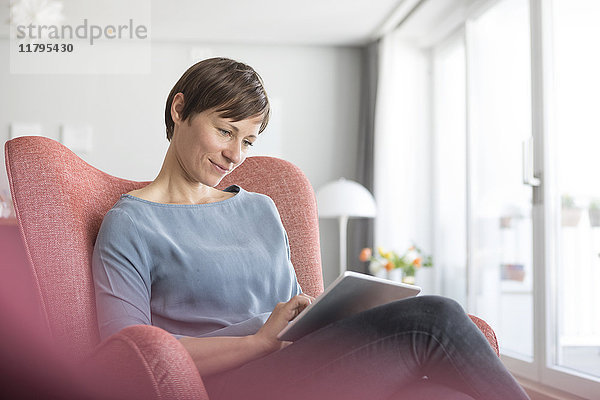 Porträt einer Frau  die zu Hause auf einem Sessel sitzt und eine Tablette benutzt.