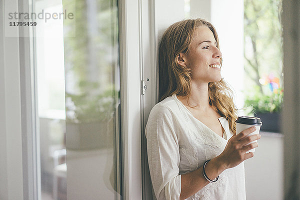 Lächelnde junge Frau mit Kaffee zum Mitnehmen am Fenster
