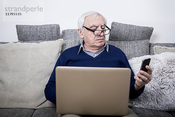 Senior Mann sitzt auf der Couch  mit Laptop und Smartphone