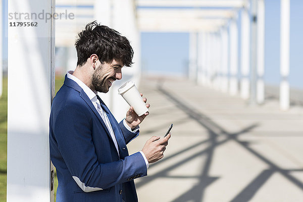 Glücklicher junger Geschäftsmann mit Smartphone und Kaffee zum Mitnehmen im Freien
