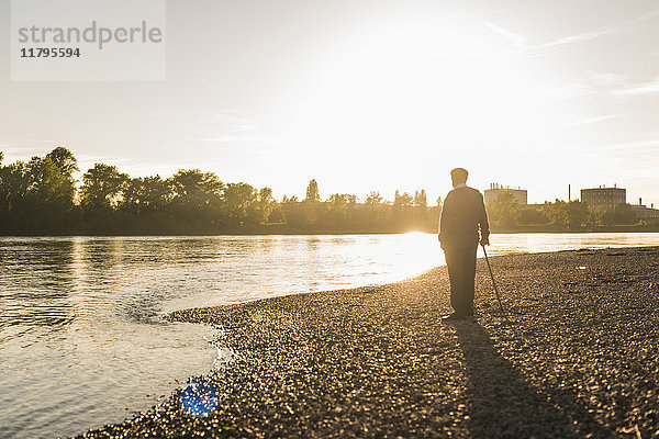 Rückansicht eines älteren Mannes  der am Flussufer steht und den Sonnenuntergang beobachtet.