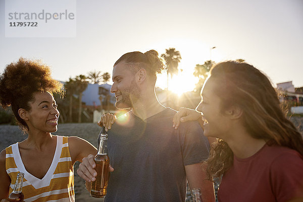 Drei Freunde mit Bierflaschen am Strand bei Sonnenuntergang