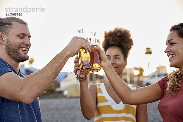 Drei Freunde stoßen mit Bierflaschen am Strand bei Sonnenuntergang an.