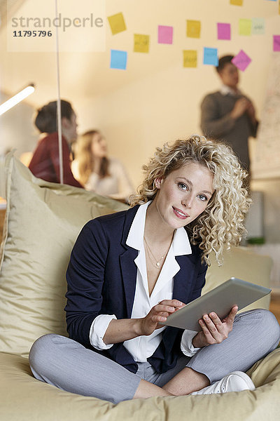 Porträt der Frau im Büro mit Tablette im Bohnenbeutel mit Besprechung im Hintergrund