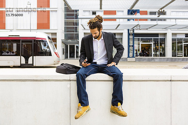 Junger Geschäftsmann mit Dreadlocks mit Smartphone beim Warten am Bahnhof