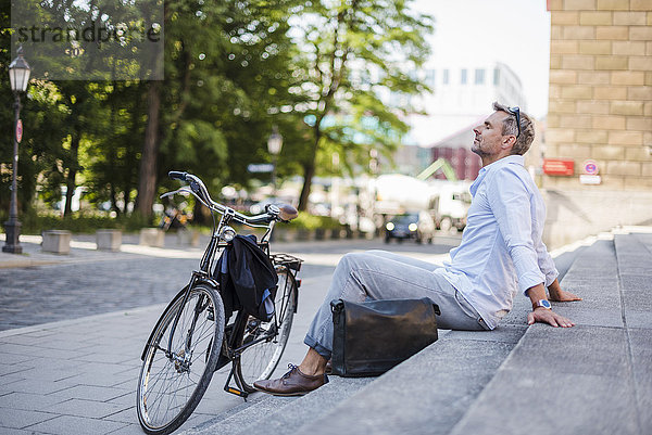 Entspannter Mann sitzt auf einer Treppe in der Stadt neben dem Fahrrad.