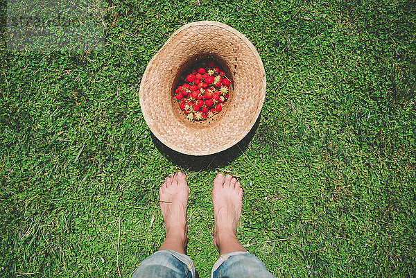 Barfüßige Frau steht neben einem Strohhut voller Erdbeeren auf dem Rasen.