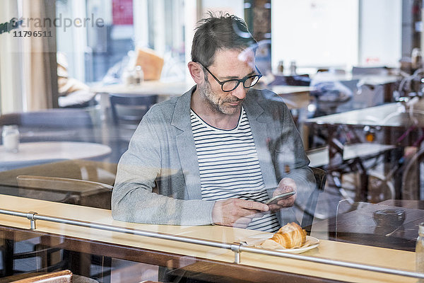 Erwachsener Mann  der hinter der Fensterscheibe eines Cafés sitzt und auf das Handy schaut.