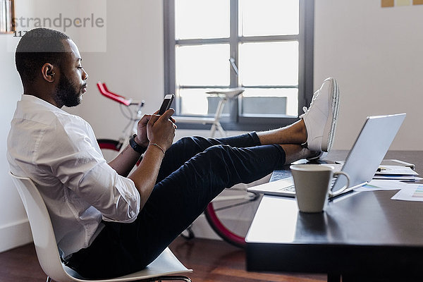 Mann mit Handy im Home-Office mit Füßen auf dem Schreibtisch