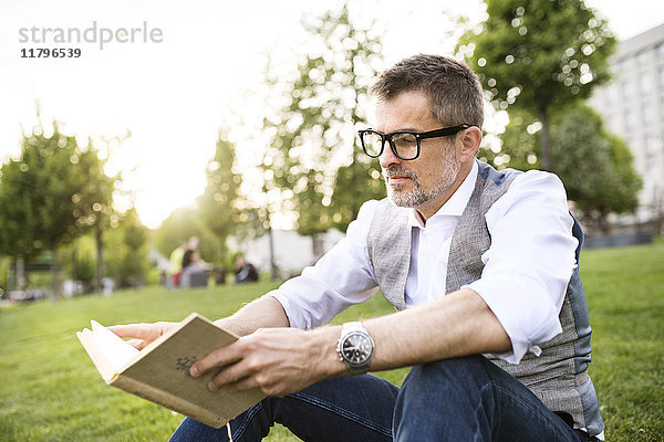 Zuversichtlicher reifer Geschäftsmann im Stadtpark auf Gras sitzend Lesebuch