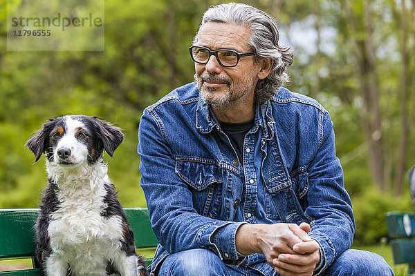 Porträt eines Mannes mit grauem Haar und Bart  der neben seinem Hund auf einer Bank sitzt.