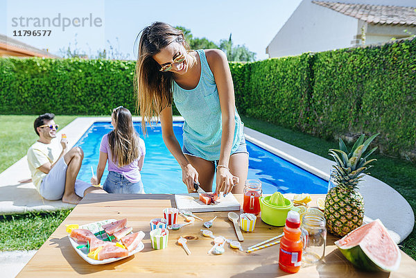 Frau beim Zubereiten von Wassermelonensaft am Pool mit Freunden im Hintergrund
