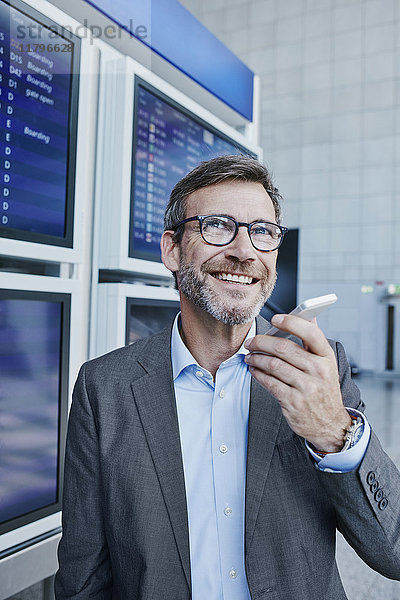 Lächelnder Geschäftsmann mit Handy zum Flugplan am Flughafen