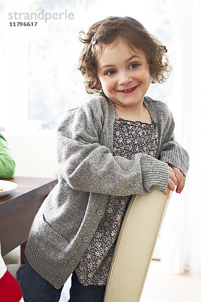 Porträt eines lächelnden kleinen Mädchens  das zu Hause auf einem Stuhl kniend sitzt