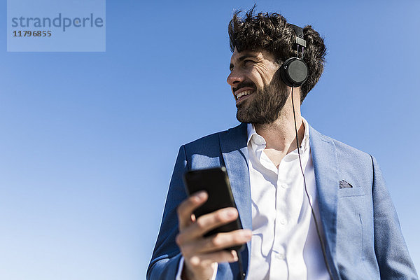 Junger Geschäftsmann mit Smartphone mit Kopfhörer unter blauem Himmel