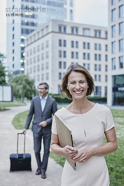 Portrait der lächelnden Geschäftsfrau im Freien mit Laptop und Geschäftsmann im Hintergrund