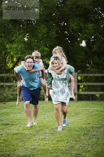 Mann und Frau rennen über einen Rasen  tragen Junge und Mädchen huckepack  eine Familie im Freien.