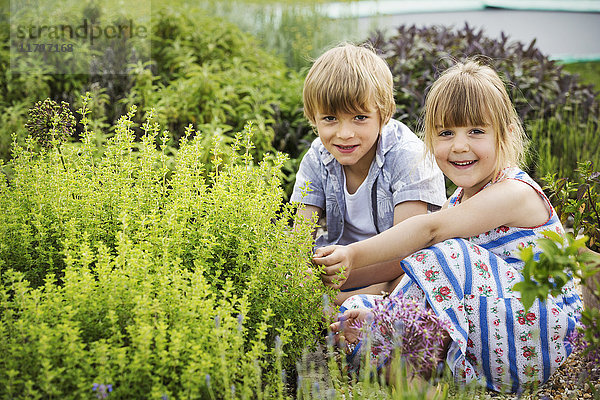 Junge und Mädchen knien nebeneinander an einem Strauch in einem Garten und lächeln in die Kamera.