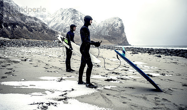 Zwei Surfer in Neoprenanzügen und mit Surfbrettern  die an einem Strand mit Bergen im Rücken stehen.