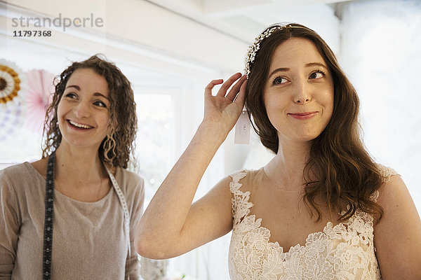 Eine junge Frau probiert Haarschmuck und Schleier mit weißer Spitze über dem Brautkleid an  unterstützt von einer Einzelhandelsberaterin.