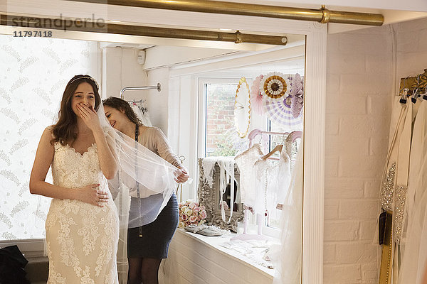 Eine Braut  eine junge Frau  die ein Hochzeitskleid mit Spitzenüberzug und Schleier anprobiert  die Hand vor Freude und Überraschung im Gesicht. Im Hintergrund eine Verkäuferin.