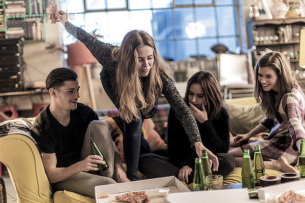 Drei junge Frauen und ein junger Mann sitzen lächelnd auf einem Sofa  Pizza und Bierflaschen auf dem Couchtisch.