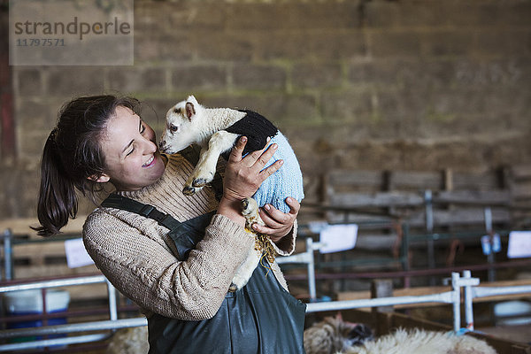 Frau steht in einer Scheune und hält ein neugeborenes Lamm in einem gestrickten Pullover in der Hand.