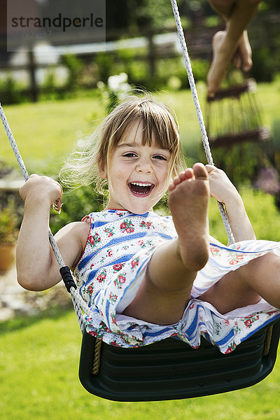Lächelndes Mädchen im Sonnenkleid auf einer Schaukel in einem Garten.