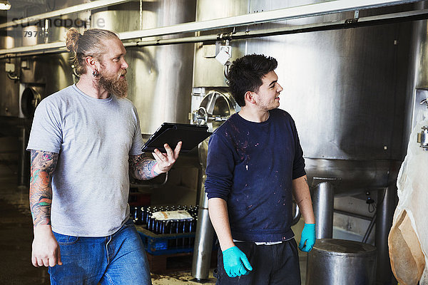 Zwei Männer  die in einer Brauerei arbeiten  gehen an großen Metallkesseln vorbei und halten ein digitales Tablett in der Hand.