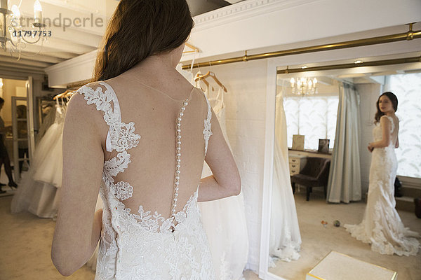 Eine junge Frau in einem weißen Hochzeitskleid in voller Länge  die ihr Spiegelbild im Spiegel in einer Brautboutique betrachtet.