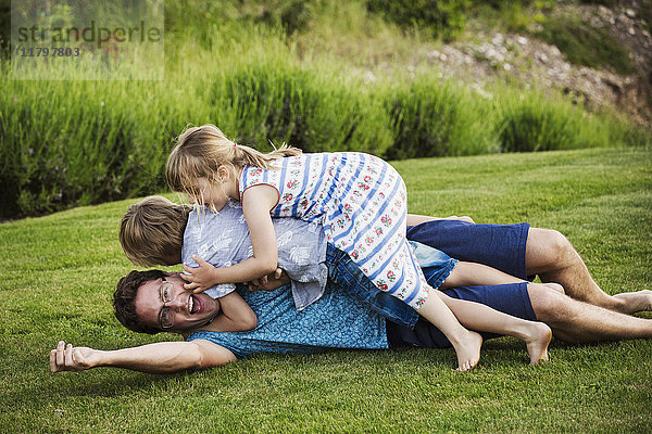 Ein Mann  der im Gras liegt und auf dem seine beiden Kinder liegen und spielen.