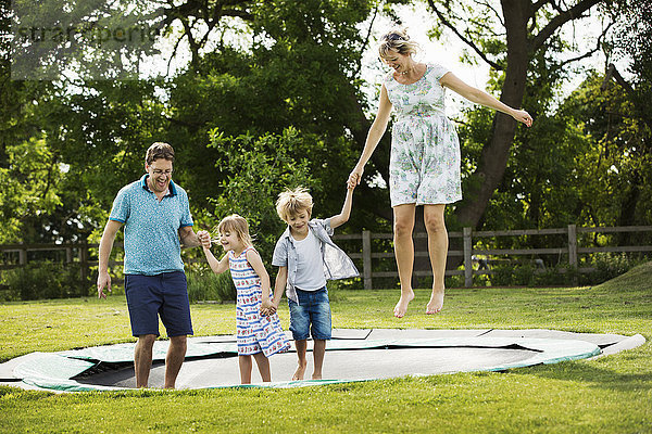 Mann  Frau  Junge und Mädchen halten sich an den Händen und springen auf einem Trampolin  das auf dem Rasen in einem Garten aufgestellt ist.