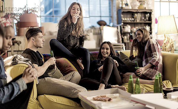 Vier junge Frauen und ein junger Mann sitzen lächelnd auf einem Sofa  Pizza und Bierflaschen auf dem Couchtisch.