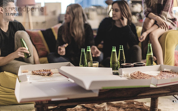 Drei junge Frauen und ein junger Mann sitzen lächelnd auf einem Sofa  Pizza und Bierflaschen auf dem Couchtisch.