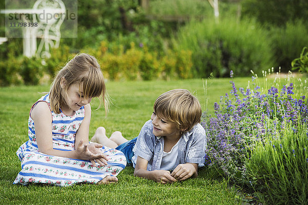 Ein Mädchen sitzt auf dem Rasen und unterhält sich mit ihrem Bruder  der neben ihr auf einem Rasen in einem Garten liegt.