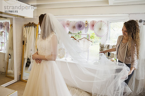 Eine junge Frau in einem weißen Hochzeitskleid in voller Länge und eine Assistentin oder Schneiderin  die ihren langen Netzschleier hochhält.