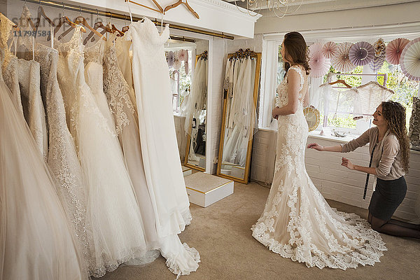 Ausgestellte Reihen von Brautkleidern. Eine junge Frau in einem weißen Hochzeitskleid in voller Länge  die ihr Spiegelbild im Spiegel in einer Brautboutique betrachtet.