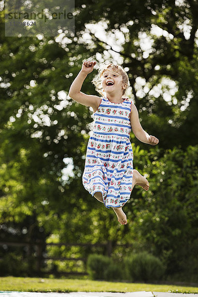 Mädchen im Sonnenkleid  das in einem Garten auf ein Trampolin springt.