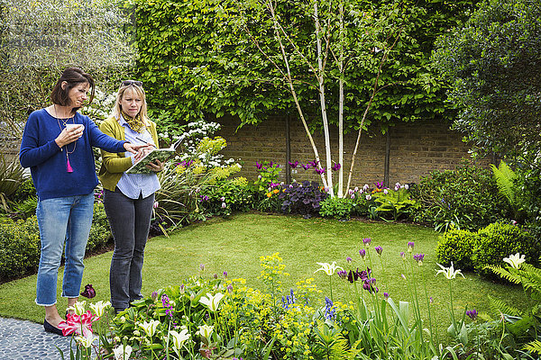 Zwei Frauen stehen in einem Garten auf einer von Blumenbeeten umgebenen Wiese und diskutieren über die Gartengestaltung.