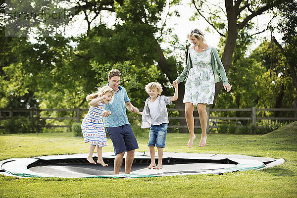 Mann  Frau  Junge und Mädchen halten sich an den Händen und springen auf einem in den Boden eingelassenen Trampolin in einem Garten.