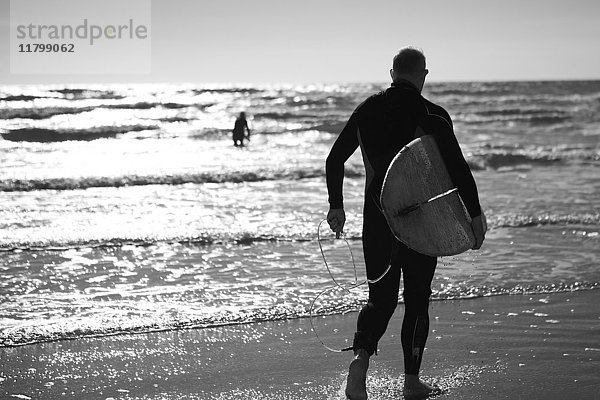 Rückansicht eines Mannes im Taucheranzug  der am Meer steht und ein Surfbrett trägt.