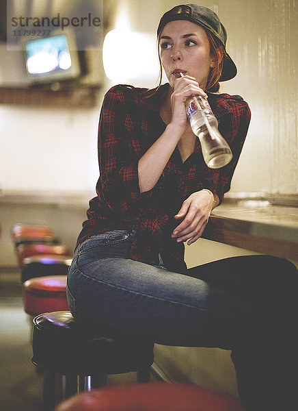 Eine Frau sitzt auf einem Hocker  trinkt in einer Bar aus einer Glasflasche und schaut ihr über die Schulter.