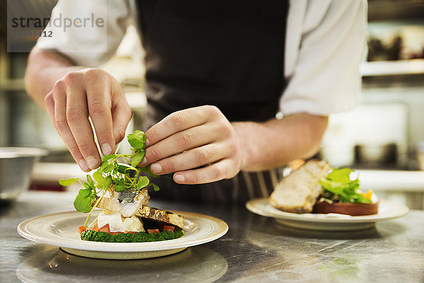 Nahaufnahme des Küchenchefs in der Küche beim Hinzufügen von Salatgarnitur zu einem Teller mit gegrilltem Fisch.