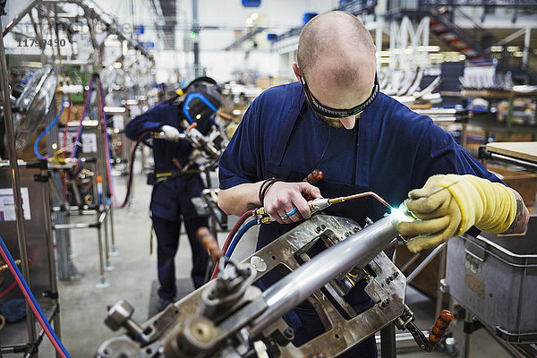 Männlicher Fabrikfacharbeiter mit Schutzbrille  der ein Schweißwerkzeug benutzt und in einer Fabrik auf einem Fahrrad arbeitet.