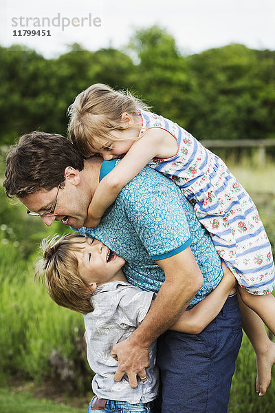 Ein Vater und zwei Kinder in einem Garten  ein Junge umarmt ihn um die Taille und ein Mädchen auf dem Rücken mit Armen um den Hals.